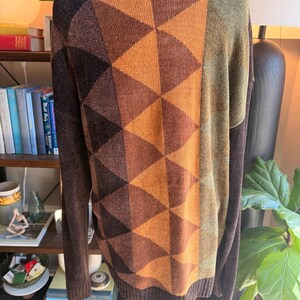 May include: A long-sleeve knit sweater featuring a geometric pattern of triangles in shades of brown, orange, and mustard yellow. The garment has a dark brown ribbed crew neckline, cuffs, and hem, with a solid olive green panel on the right side.