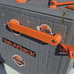 May include: A gray storage crate with a grid-patterned exterior, featuring bright orange handles and bungee cord attachments. The side displays the black and orange YakAttack logo. A small sticker depicting a person fishing is placed near the handle.