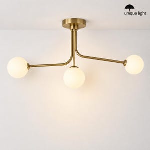 May include: A ceiling light fixture featuring a central brass-toned metal mount with three extending arms. Each arm terminates in a spherical, frosted white glass globe. The fixture includes a small logo with an umbrella icon and the text unique light.