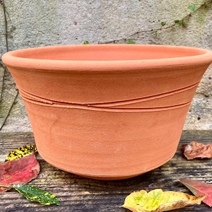 Puede incluir: Una maceta de terracota con un borde ancho y redondeado y un acabado mate de color naranja terroso. La superficie exterior presenta dos líneas incisas horizontales, distintas y ligeramente irregulares, que rodean la parte superior del cuerpo.