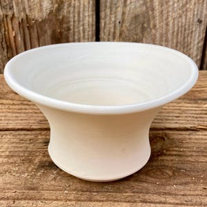 May include: A white ceramic bowl with a wide, flared rim and a tapered base. The bowl features a smooth, matte finish with subtle horizontal texture lines visible along the interior and exterior surfaces, creating a simple, functional vessel.
