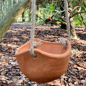 Op de afbeelding: Een hangende plantenbak van terracotta-kleurige klei met een ronde, komvormige vorm en een onregelmatige, golvende rand. De plantenbak hangt aan twee dikke, lichtgekleurde gedraaide jutekoorden die door gaten aan weerszijden zijn geknoopt.