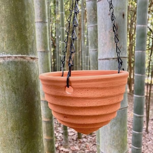 Pode incluir: Um vaso suspenso de terracota com formato cónico e nervurado. O vaso apresenta um acabamento mate em castanho-alaranjado e é suspenso por três correntes de metal preto fixadas a pequenos orifícios ao longo do rebordo superior do vaso.