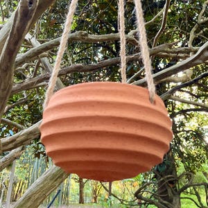 May include: A round, terracotta-colored hanging planter with a ribbed, horizontal texture. The planter features a wide, bowl-like shape and is suspended by two thick, natural-colored jute ropes attached to the sides of the rim.