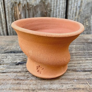 May include: A small, terracotta-colored clay pot with a wide, flared rim and a textured, slightly uneven surface. The vessel features a rounded, bulbous body that tapers toward a narrow base, with a small, stamped maker's mark visible on the side.