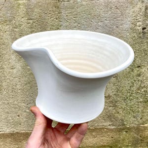 May include: A white ceramic vessel with a smooth, glossy finish and a distinctive asymmetrical, curved rim. The bowl features a wide, flared opening that tapers down to a narrower base, showcasing subtle horizontal wheel-thrown texture lines.