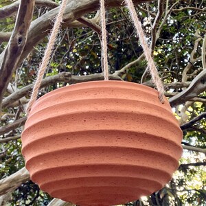 Peut inclure: Un pot de fleurs suspendu de couleur terre cuite, de forme ronde, présentant une série de rainures horizontales et nervurées sur sa surface extérieure. Le récipient en forme de bol est suspendu par trois cordes en jute naturel.