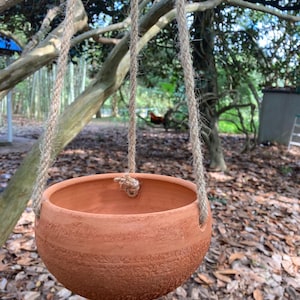 May include: A round terracotta hanging planter with a smooth upper rim and a textured, rough lower body. The bowl is suspended by three thick, natural-colored jute ropes that pass through holes in the sides and are knotted at the top.