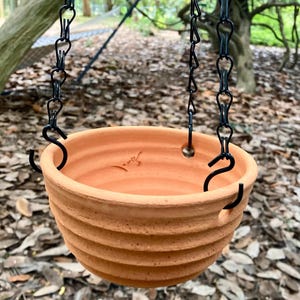 Può includere: Un vaso da fiori sospeso color terracotta con texture a coste orizzontali sull'esterno. La ciotola presenta tre fori equidistanti vicino al bordo, ciascuno collegato a una catena di metallo nero che converge verso l'alto per la sospensione.