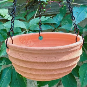May include: A round, terracotta-colored hanging planter with a ribbed, textured exterior surface. The bowl-shaped vessel features three evenly spaced holes along the upper rim, which are attached to a black metal chain for suspension purposes.