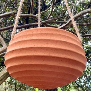 Peut inclure: Un pot de fleurs suspendu de couleur terre cuite, de forme ronde avec une texture nervurée horizontale. Le récipient en forme de bol est suspendu par trois brins de corde de jute de couleur naturelle fixés au bord supérieur.