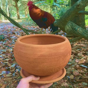 May include: A round, terracotta-colored clay planter pot with a matching circular saucer base. The pot features a textured, matte surface with visible horizontal striations and a wide, open top rim, designed for holding plants or decorative items.
