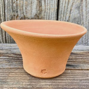 May include: A terracotta planter with a wide, flared rim and a tapered body. The surface of the clay pot is matte and light orange in color. A small, stamped maker's mark is visible near the base of the exterior wall of the vessel.