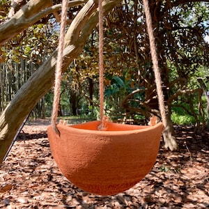 Puede incluir: Una maceta colgante de terracota con diseño en forma de cuenco y textura rugosa y porosa. La maceta está suspendida por tres cuerdas de yute gruesas de color natural, anudadas a través de orificios en el borde superior del recipiente.