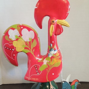 May include: A red rooster figurine with a large crest, yellow beak, and green base. The body features painted white and yellow flowers, green leaves, and heart shapes. A small tag attached to the base displays a blue rooster and the text ROOSTER D'AMOUR.