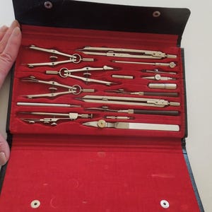 May include: A vintage drafting set in a black case with a red velvet interior. The set includes various silver-colored compasses, dividers, and other drawing tools. The case is open, revealing the neatly arranged instruments.