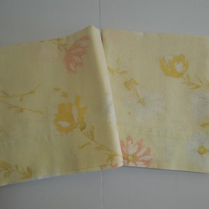 May include: Two folded yellow fabric napkins with a floral pattern. The pattern features yellow, pink, and white flowers with green stems and leaves. The napkins are likely made of a soft, woven material.