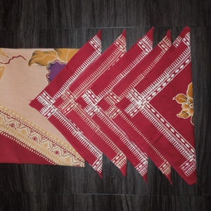 May include: A set of six red and white patterned cloth napkins, folded into triangles, next to a matching rectangular tablecloth. The tablecloth has a beige background with floral and geometric designs in red, yellow, and purple.