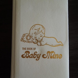 Puede incluir: Un libro color crema titulado "The Book of Baby Mine" con letras doradas y una ilustración dorada de un bebé. El bebé está acostado y sosteniendo un juguete. El libro tiene una cubierta texturizada.