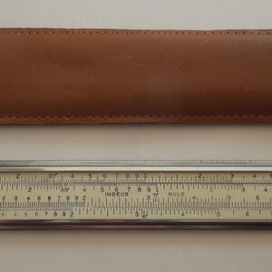 May include: A vintage slide rule with a cream-colored scale and silver metal edges, next to a brown leather case. The slide rule has various markings and numbers, and the case is rectangular.