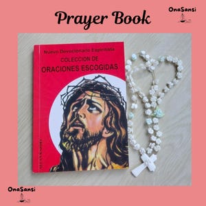 Può includere: Un libro di preghiere rosso intitolato Nuevo Devocionario Espiritista Coleccion de Oraciones Escogidas con l'illustrazione di un uomo con una corona di spine, accompagnato da un rosario bianco con perline a forma di fiore e una croce.