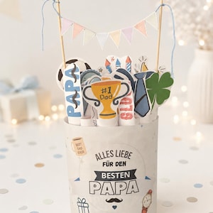 May include: A cylindrical paper container featuring the text ALLES LIEBE FÜR DEN BESTEN PAPA, decorated with icons of a mug, gift, mustache, and watch. It holds various paper cutouts including a #1 Dad trophy, a soccer ball, and a tie, topped with a bunting.