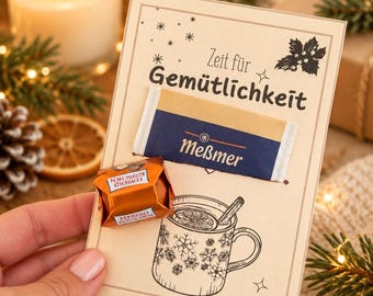 May include: A rectangular card featuring the text Zeit für Gemütlichkeit, a Meßmer tea bag, and a Ferrero Küsschen chocolate. The card includes a black ink illustration of a mug with snowflake patterns and a cinnamon stick.