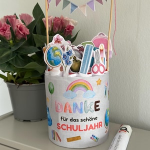 May include: A cylindrical container decorated with a rainbow and school supplies, featuring the text "DANKE für das schöne SCHULJAHR". It holds various paper cutouts of school items and is topped with a small bunting banner on two wooden sticks.