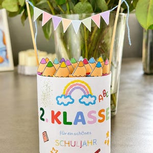 Puede incluir: Un recipiente cilíndrico blanco lleno de recortes de lápices de papel, con un arcoíris colorido, el texto 2. KLASSE y el texto für ein schönes SCHULJAHR. Dos palos de madera sostienen una pequeña guirnalda de banderines de colores pastel.