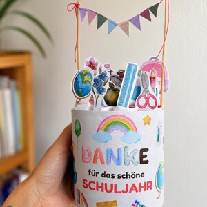 May include: A cylindrical paper container featuring a rainbow and school-themed illustrations with the text "DANKE für das schöne SCHULJAHR". The top is decorated with wooden sticks holding a colorful bunting and various school supply cutouts.