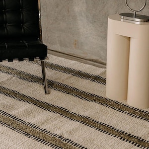 Hand woven wool area rug in cream, olive green, and black with a modern striped pattern, featuring a textured flatweave design and neutral colors, shown in close-up and contemporary living room settings. Handmade minimalist rug for living room, bedroom, office, or entryway decor.