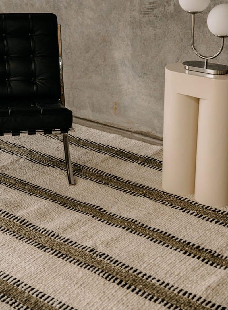 Hand woven wool area rug in cream, olive green, and black with a modern striped pattern, featuring a textured flatweave design and neutral colors, shown in close-up and contemporary living room settings. Handmade minimalist rug for living room, bedroom, office, or entryway decor.