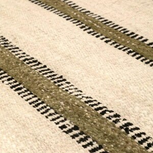 Hand woven wool area rug in cream, olive green, and black with a modern striped pattern, featuring a textured flatweave design and neutral colors, shown in close-up and contemporary living room settings. Handmade minimalist rug for living room, bedroom, office, or entryway decor.
