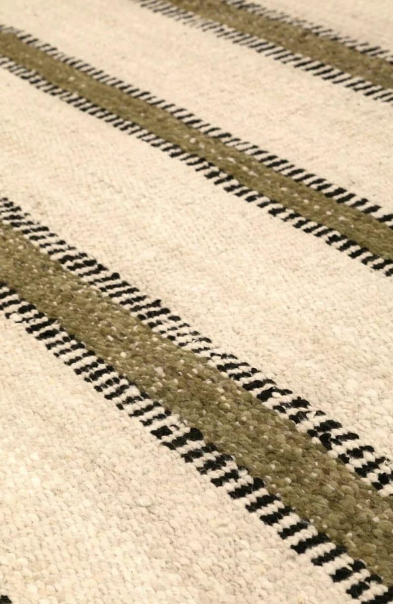 Hand woven wool area rug in cream, olive green, and black with a modern striped pattern, featuring a textured flatweave design and neutral colors, shown in close-up and contemporary living room settings. Handmade minimalist rug for living room, bedroom, office, or entryway decor.