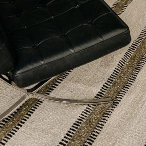 Hand woven wool area rug in cream, olive green, and black with a modern striped pattern, featuring a textured flatweave design and neutral colors, shown in close-up and contemporary living room settings. Handmade minimalist rug for living room, bedroom, office, or entryway decor.