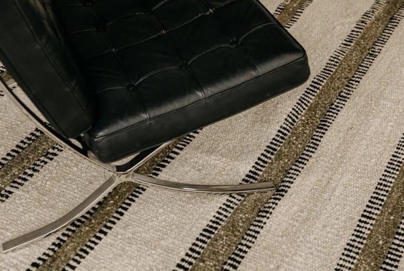Hand woven wool area rug in cream, olive green, and black with a modern striped pattern, featuring a textured flatweave design and neutral colors, shown in close-up and contemporary living room settings. Handmade minimalist rug for living room, bedroom, office, or entryway decor.