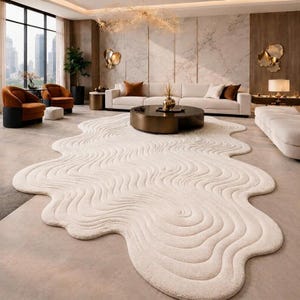 May include: An off-white area rug with an irregular, organic shape and a textured surface featuring concentric, wavy, topographical lines. The rug is positioned on a smooth floor in a room with a sofa, armchairs, and a round coffee table.
