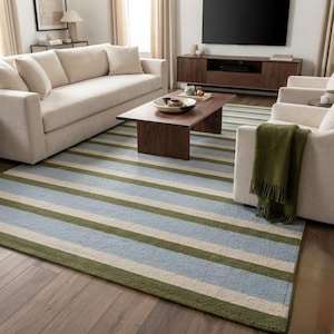 May include: A rectangular area rug featuring a repeating horizontal striped pattern in shades of light blue, olive green, and cream. The rug is positioned on a dark wood floor, extending underneath a beige sofa and a wooden coffee table.