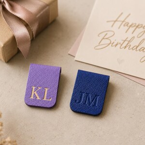 Op de afbeelding: Twee rechthoekige magnetische boekenleggers met een textuurafwerking. De ene is paars met de goudkleurige letters KL in reliëf, de andere is donkerblauw met de letters JM in reliëf. Beide items hebben een vouwbaar ontwerp om pagina's vast te zetten.
