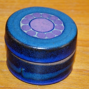 Op de afbeelding: Een kleine, ronde keramische pot met deksel, voorzien van een diepblauw glazuur met een donkere, bijna zwarte basis. Het deksel is versierd met een centrale paarse cirkel omringd door een ring van kleinere paarse stippen op een lichtgrijze achtergrond.
