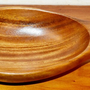 Puede incluir: Un cuenco de madera con forma de pez con un acabado suave y pulido y patrones de veta de madera natural visibles. El cuenco presenta una cuenca central ancha y redondeada que se estrecha hacia la cola y la cabeza, mostrando un tono marrón medio cálido.