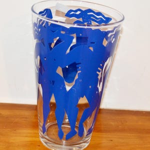 Op de afbeelding: Een helder drinkglas met een opvallende, blauwe grafische print van gestileerde figuren in beweging. Het ontwerp loopt rond de buitenkant van het glas en toont abstracte menselijke vormen in een levendige kobaltblauwe kleur op het glas.