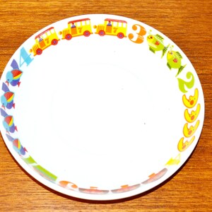 Könnte beinhalten: Eine weiße, runde Schüssel mit einem farbenfrohen Randdesign. Das Muster zeigt gelbe Busse mit der Nummer 7, grüne Fische, blaue Vögel und Mondsicheln, durchsetzt mit den Zahlen 4, 3 und 2, die in leuchtenden, kräftigen und bunten Schriftarten gedruckt sind.