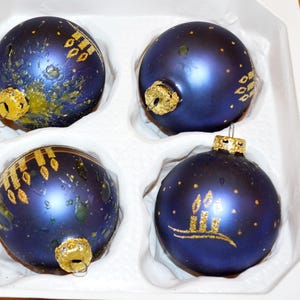 Puede incluir: Un conjunto de cuatro adornos esféricos de vidrio azul oscuro con diseños de velas con purpurina dorada y tapas de metal dorado. Cada adorno presenta un acabado mate suave con detalles dorados pintados a mano, dispuestos en una bandeja blanca.