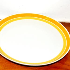Op de afbeelding: Een rond wit keramieken dinerbord met een brede, felgele band rondom de rand. Het bord heeft een gladde, glanzende afwerking en een strak, eenvoudig ontwerp met een effen wit midden, wat zorgt voor een helder en functioneel stuk serviesgoed.