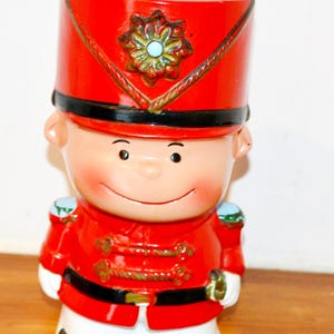 May include: A ceramic figurine of a soldier wearing a bright red uniform with a tall red hat, black belt, and white trousers. The soldier has a smiling face, rosy cheeks, and a gold-toned emblem on the front of the hat with a small blue center.
