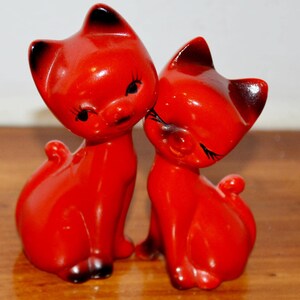 Puede incluir: Dos figuras de gato de cerámica roja brillante colocadas una al lado de la otra. Cada gato presenta orejas puntiagudas con puntas negras, ojos negros pintados con pestañas largas y una pequeña cola rizada. Los gatos tienen superficies lisas.