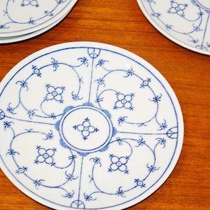 Könnte beinhalten: Ein Set aus weißen Keramiktellern mit blauem Unterglasur-Dekor. Jeder Teller zeigt ein zentrales kreisförmiges Motiv mit einer vierblättrigen Blume, umgeben von symmetrischen Rankenmustern und weiteren floralen Elementen in blauer Farbe.