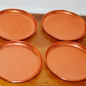Puede incluir: Un conjunto de cuatro platos redondos de color cobre metálico dispuestos en una cuadrícula de dos por dos. Cada plato presenta una textura fina y repetitiva de cuadros en toda su superficie, creando un aspecto uniforme y reflectante.