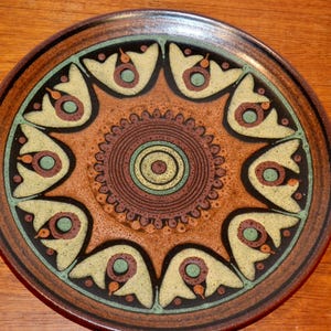 Puede incluir: Un plato de cerámica redondo que presenta un motivo circular central con anillos concéntricos en tonos marrón, verde y crema. El borde exterior muestra un patrón repetitivo de diez formas de pétalos estilizados de color crema con acentos.